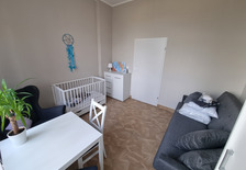 Zdjęcie pokazujące wyposażenie pokoju w Domu dla matek z małoletnimi dziećmi i kobiet w ciąży w Dobieszczyźnie. Od prawej szara kanapa, na której znajdują się poduszki, szafka stojąca w rogu pokoju, na której znajdują się lampka oraz obraze