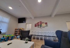 Zdjęcie pokazujące wyposażenie wspólnego pokoju w Domu dla matek z małoletnimi dziećmi i kobiet w ciąży w Dobieszczyźnie.  Z prawej strony na pierwszym planie znajduje się fotel, przy którym stoi stolik. Na drugim planie pod ścianą stoi rząd